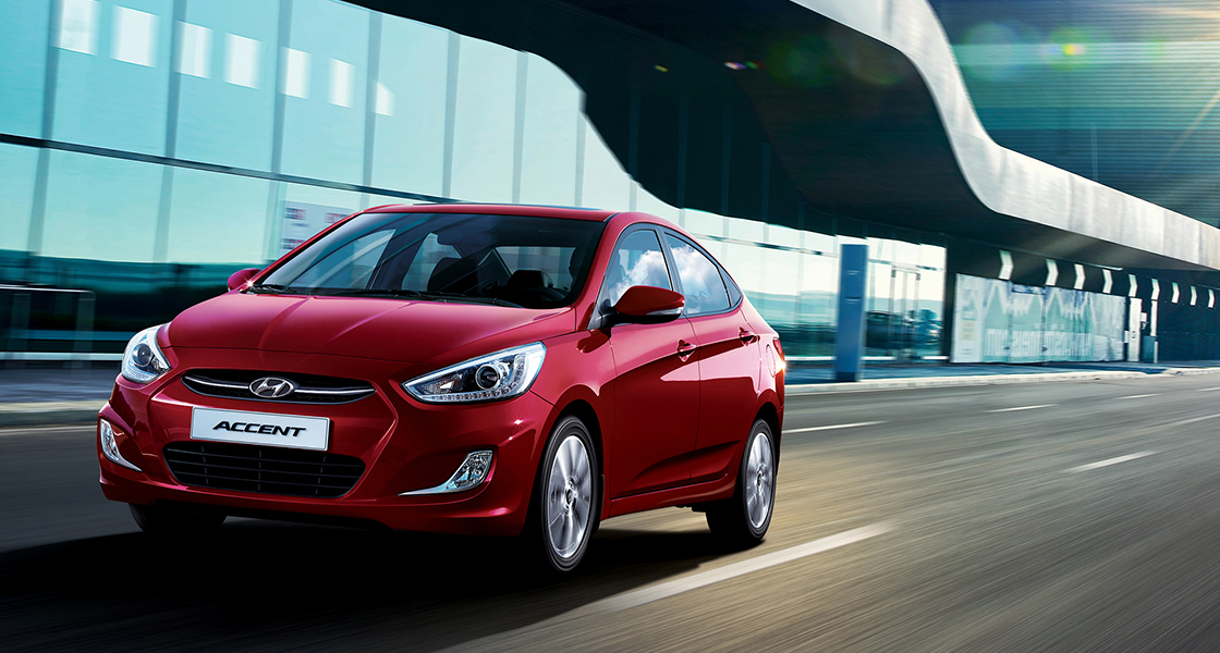 منظر أمامي جانبي من اليسار لسيارة أكسنت حمراء اللون تمر بجانب مبنى مكون من طابق واحد