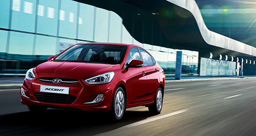 منظر أمامي جانبي من اليسار لسيارة أكسنت حمراء اللون تمر بجانب مبنى مكون من طابق واحد