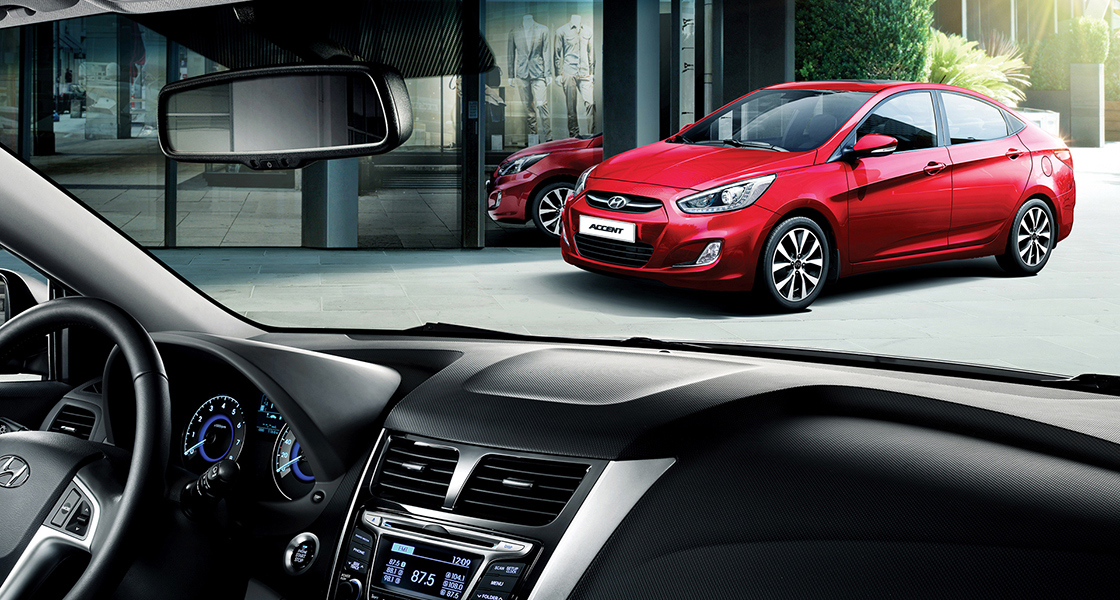 منظر أمامي جانبي لسيارة أكسنت الحمراء متوقفة أمام مبنى من زاوية نظر الراكب عبر الزجاج الأمامي