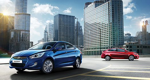 منظر أمامي جانبي لسيارة أكسنت الزرقاء في الأمام ومنظر جانبي لسيارة أكسنت الحمراء متوقفة في موقف للسيارات مع منظر للمدينة في الخلفية في يومٍ صاف