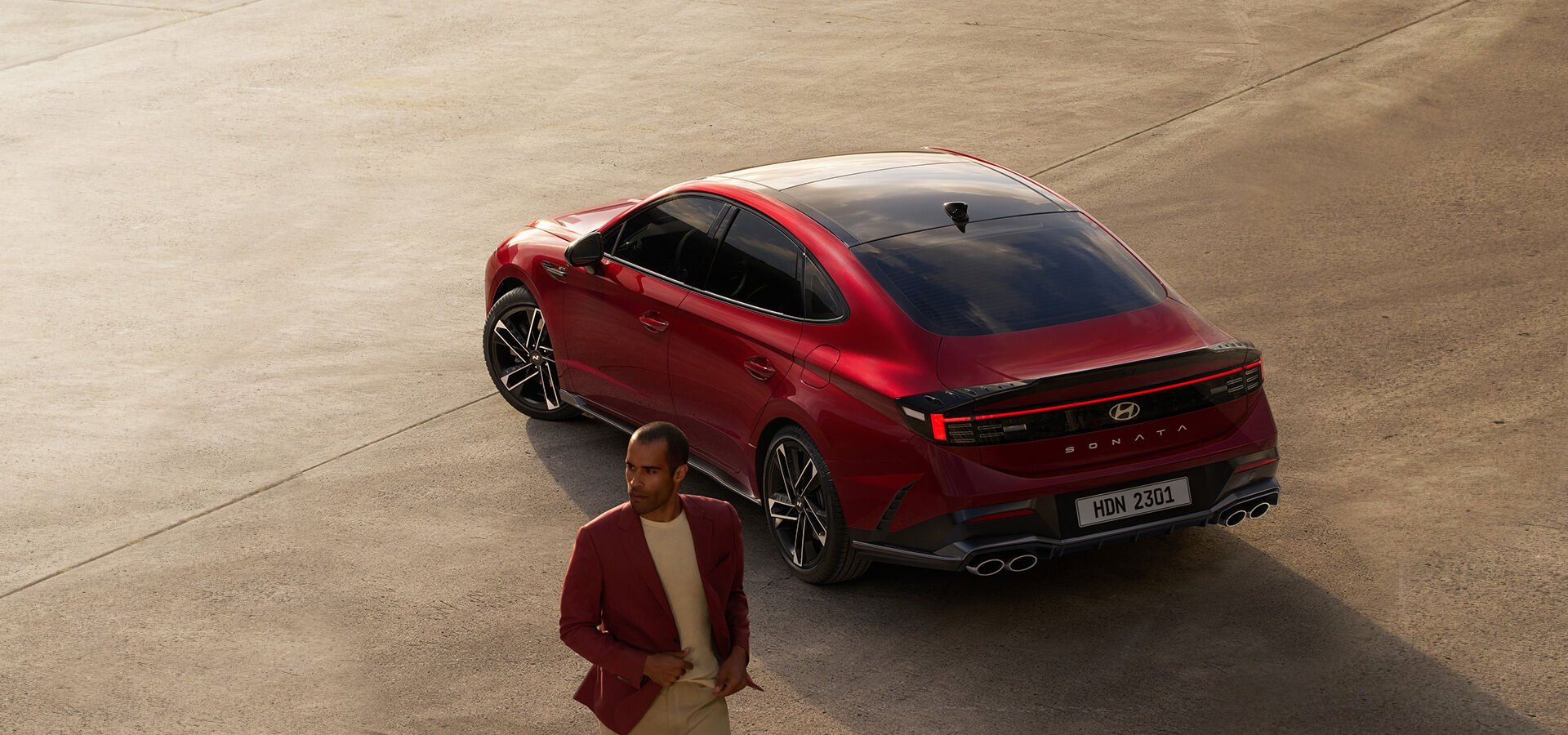 The back of the left side of the red SONATA is visible and a man in a red jacket is standing in front of the SONATA with the SONATA behind.