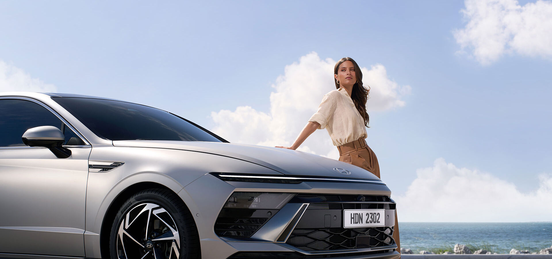 The right side of the gray SONATA is visible, and behind it a woman is leaning her arm against the SONATA. In the background, you can see the sea and the blue sky with some clouds.