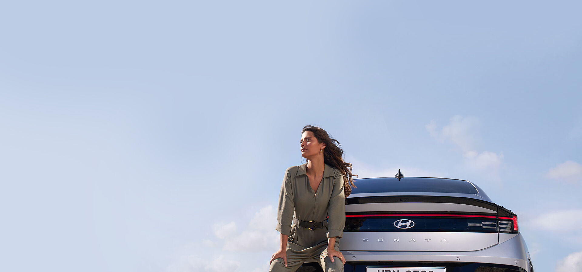 A bottom-up view of the back of the gray SONATA. A woman stands in front of the SONATA, leaning against the SONATA and looking at the sky.